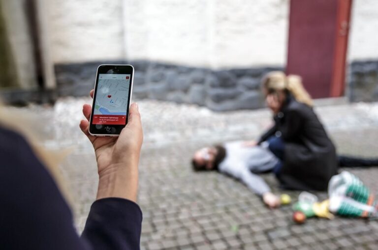 Bild på person som håller i mobil med app uppe och person som utför hjärt- och lungräddning i bakgrunden